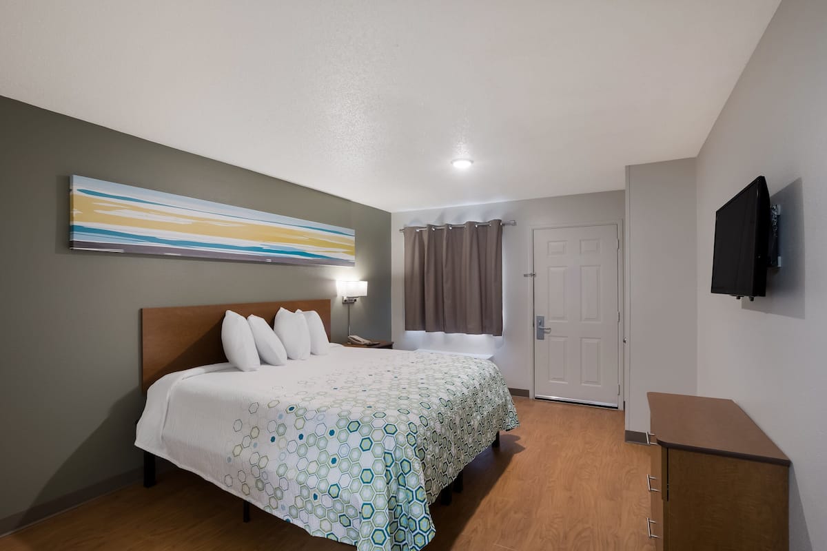 Ein Hotelzimmer mit einem Bett, einem an der Wand montierten Fernseher, einer Tür und einem Fenster mit Vorhängen.
