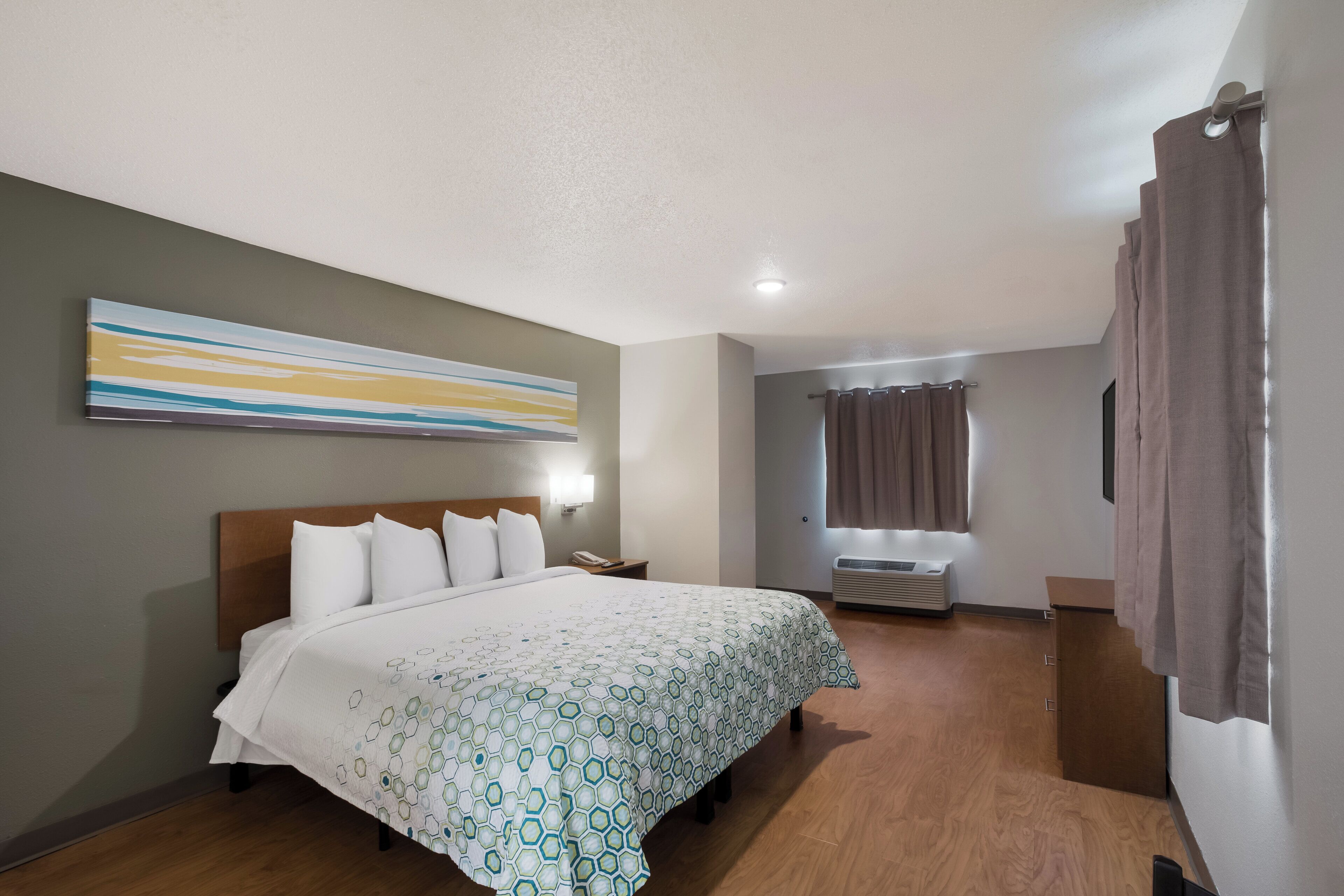 A hotel room with a bed, a nightstand, a telephone, a window with curtains, and a wall-mounted light.