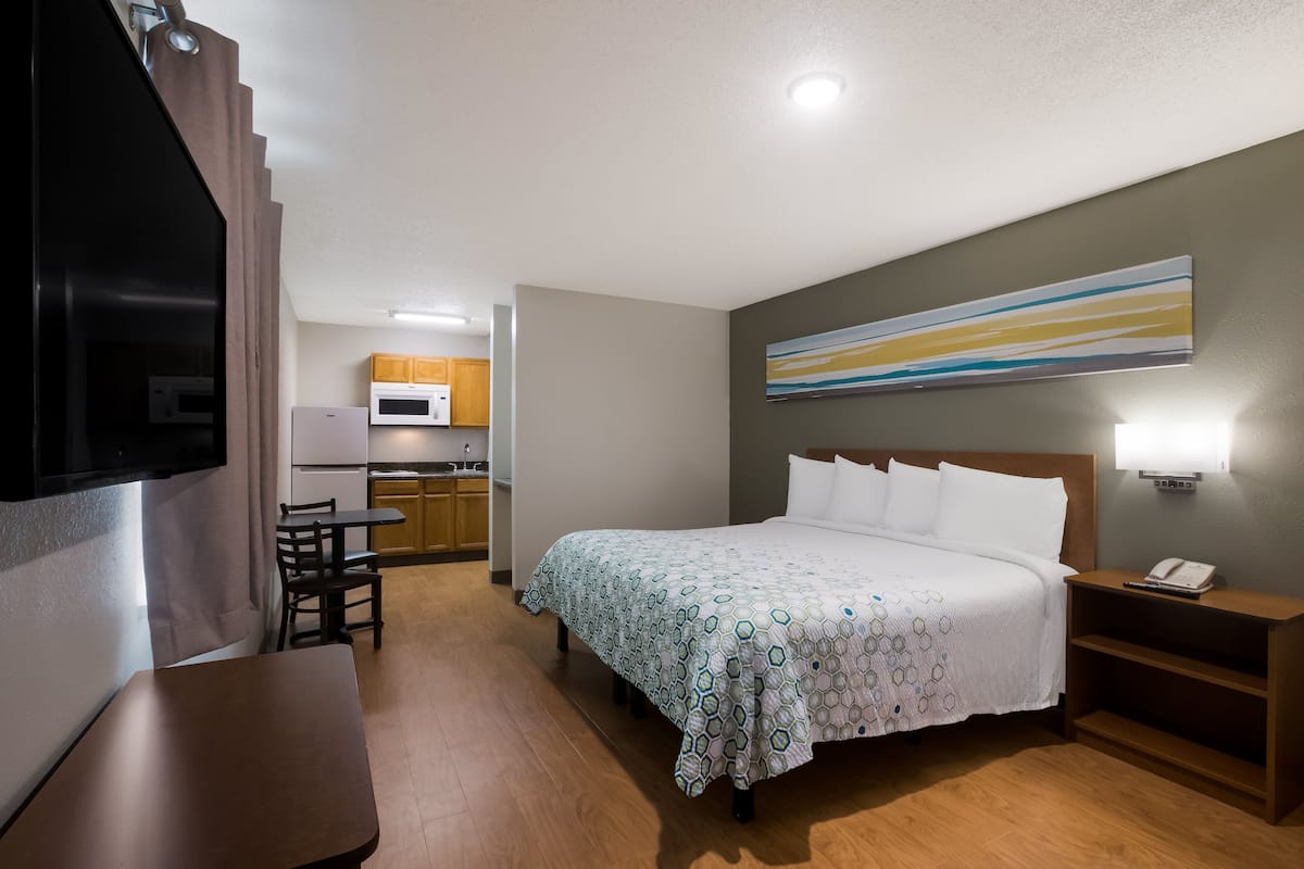 Ein Hotelzimmer mit Bett, Nachttisch und Schreibtisch mit Stühlen. Im Hintergrund ist eine Küchenzeile zu sehen.