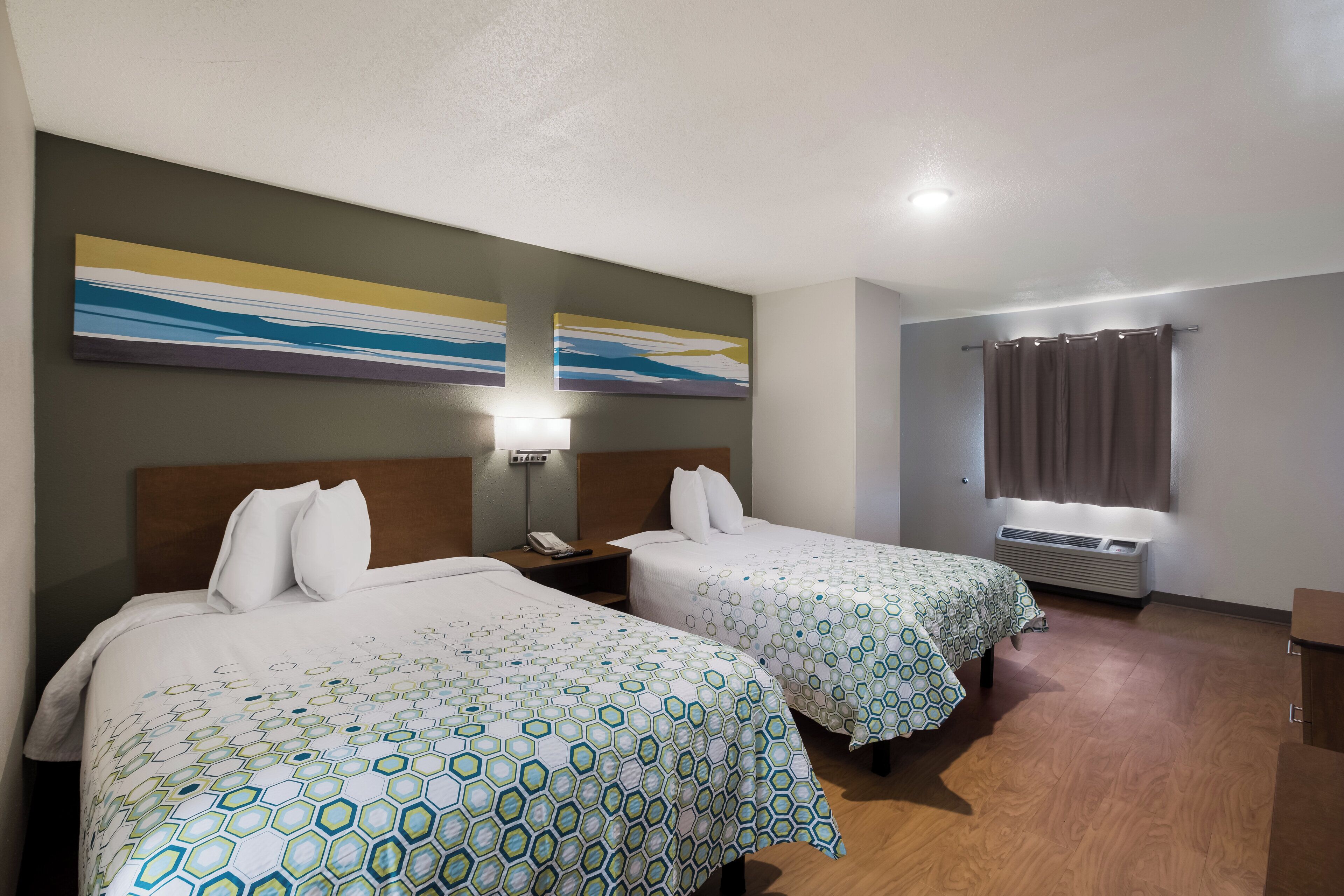 A hotel room with two beds, a nightstand, a lamp, a window with curtains, and a wall-mounted air conditioner.