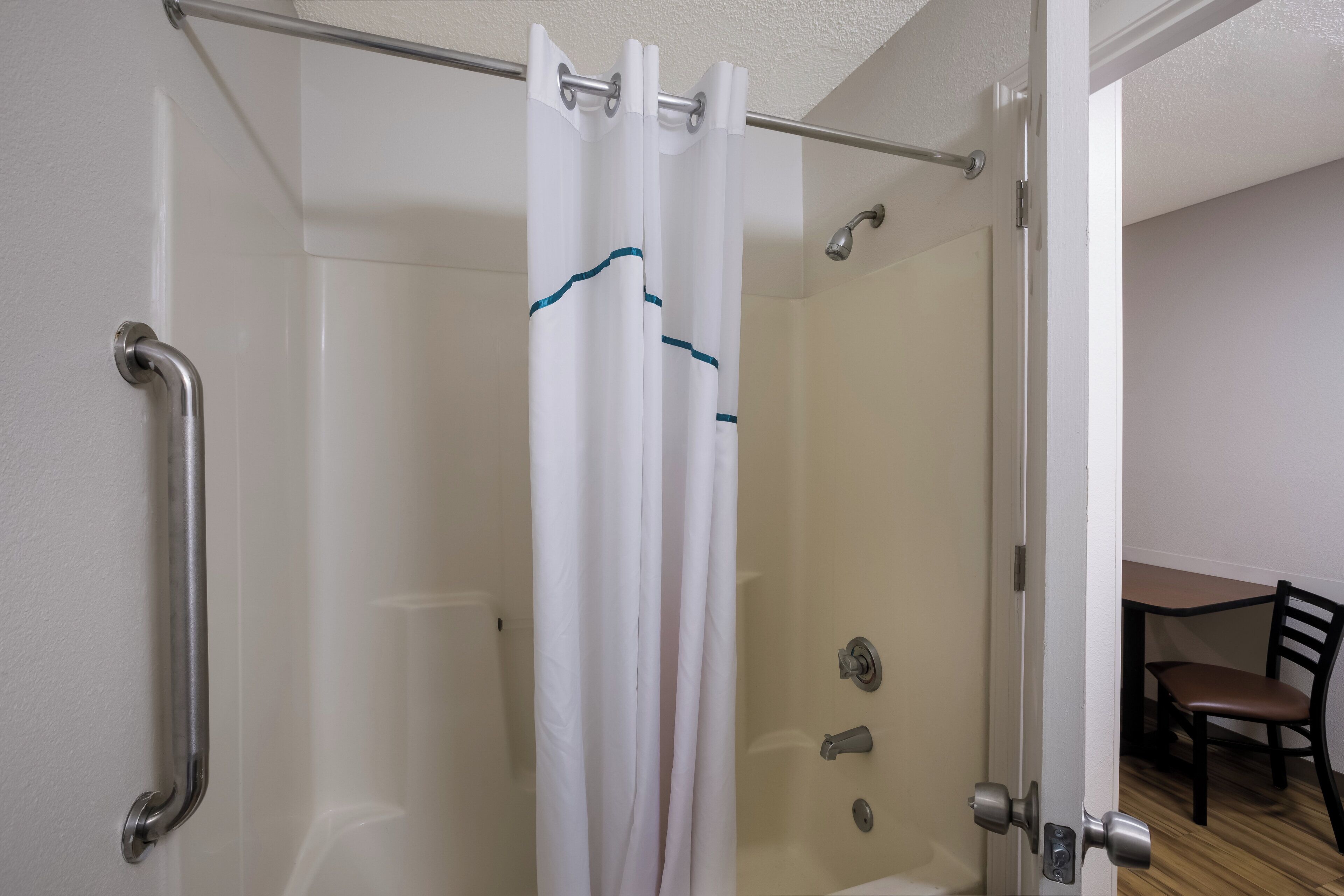 A glass-enclosed shower with a white curtain, a door with a silver handle, and a chair in the adjacent room.