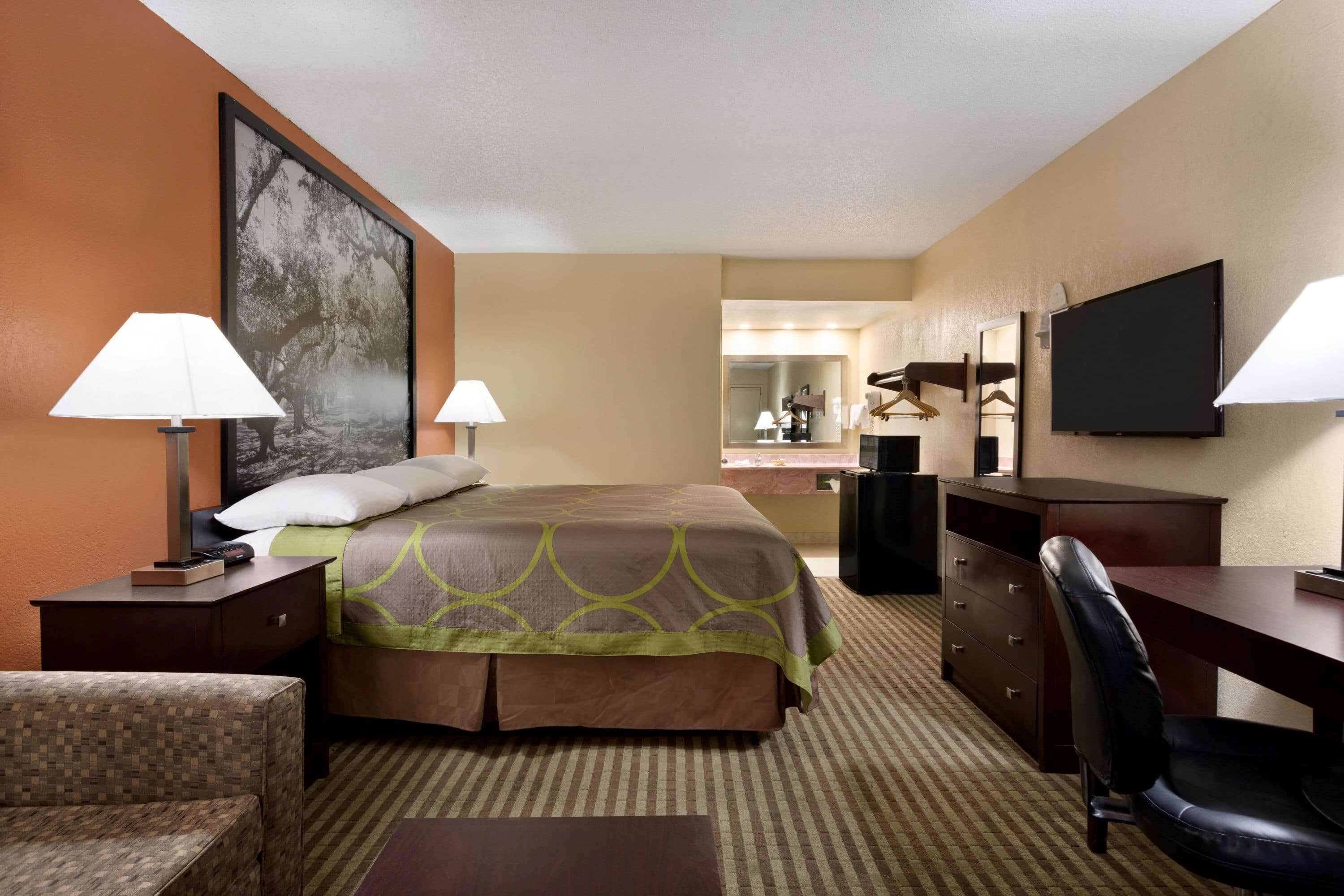 A hotel room with a bed, two bedside lamps, a desk with a chair, a television, a large mirror, and a framed picture on the wall.