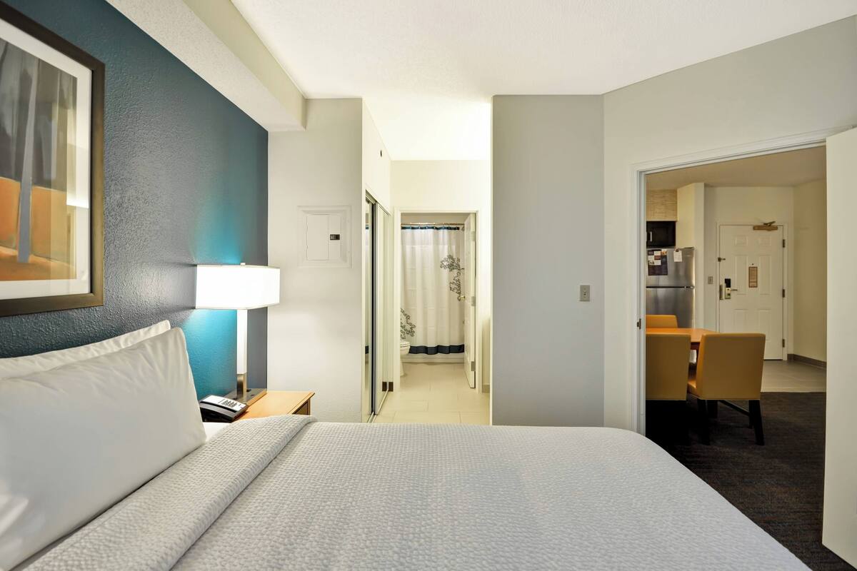 Hotellihuone, jossa on sänky, yöpöytä, lamppu, kehystetty kuva, kylpyhuone suihkulla sekä ruokailutila pöydällä ja tuoleilla.