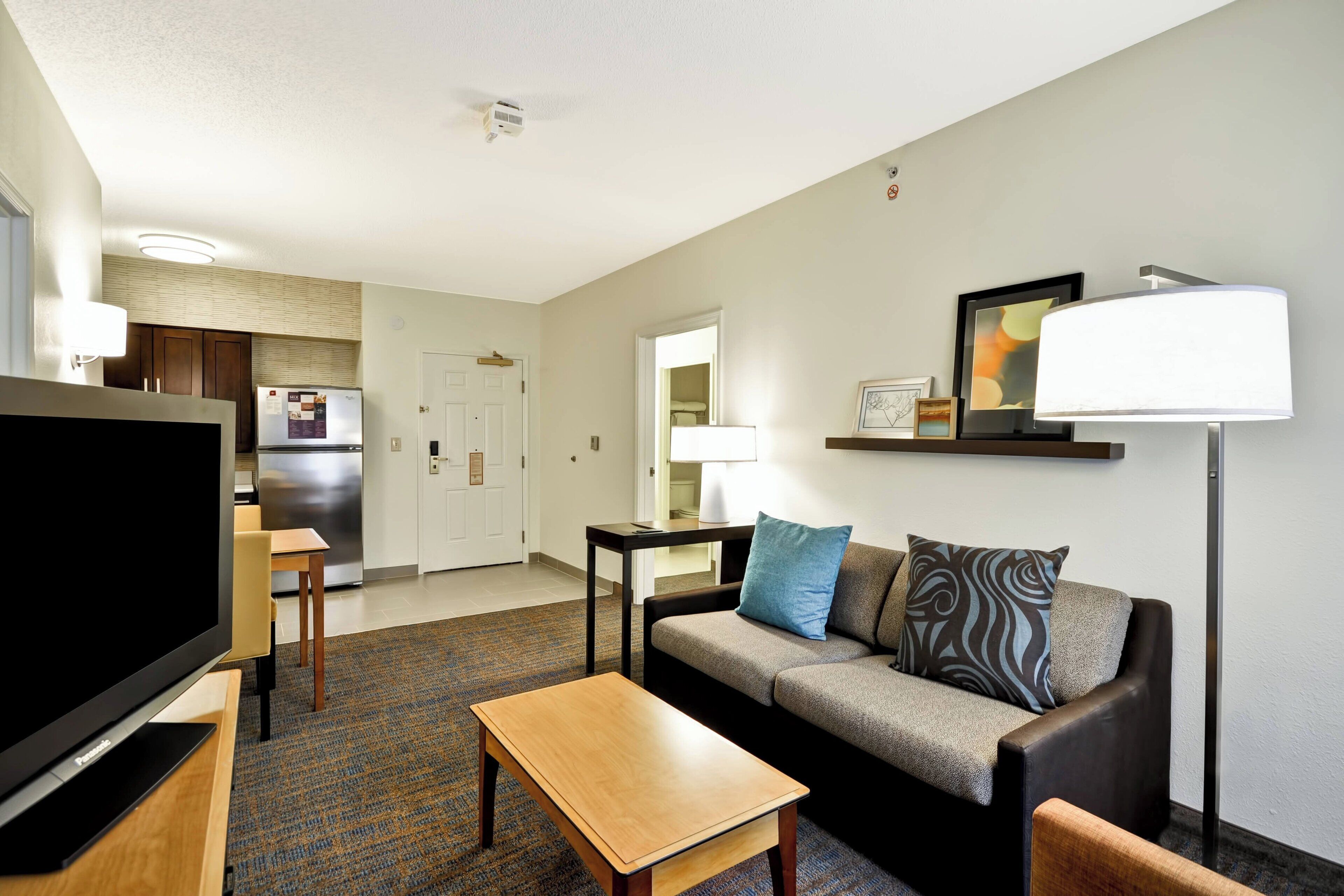 A modern hotel room with a flat-screen TV, a sofa, a coffee table, a dining area with a table and chairs, and a kitchen with a refrigerator.