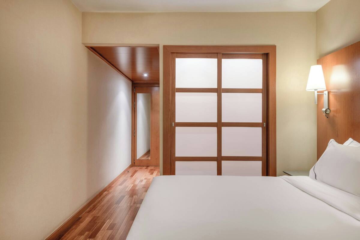 A hotel room with a wooden floor, a bed with white bedding, a nightstand, and a wall-mounted lamp.