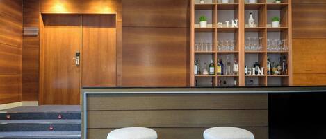 Zona de bar moderna con paneles de madera, mostrador, taburetes y estantes con vasos y botellas.