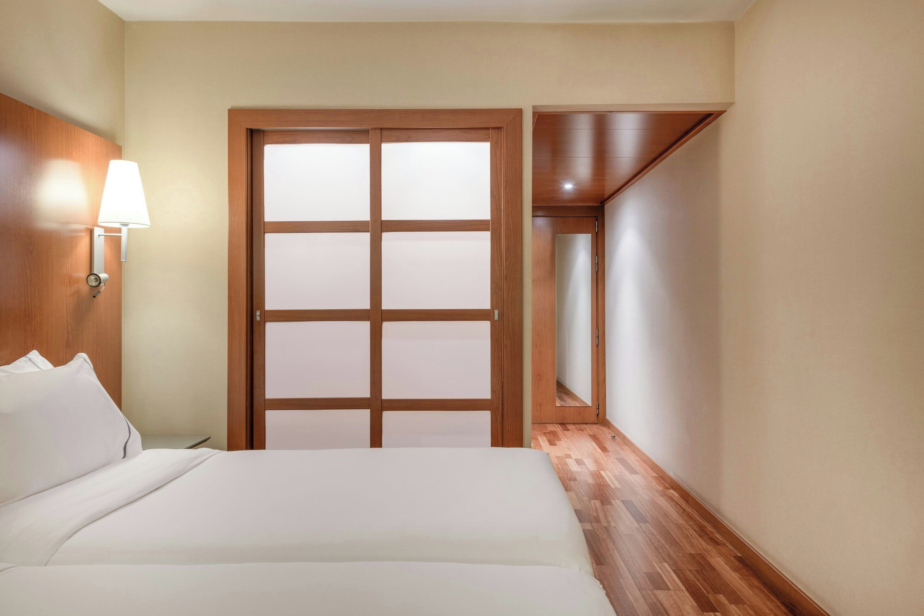 A hotel room with a bed, a nightstand, a wall-mounted lamp, and a wooden door with frosted glass panels.