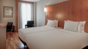 Une chambre d’hôtel avec deux lits, un bureau, une chaise, une fenêtre avec des rideaux et une lampe fixée au mur.