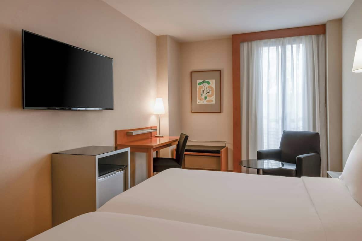 A hotel room with a large bed, a desk with a chair, a television mounted on the wall, a small table, and a window with curtains.