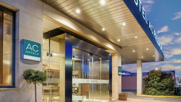 L’extérieur de l’hôtel AC présente un design moderne, avec une façade en verre et une enseigne indiquant qu’il fait partie de la chaîne Marriott.