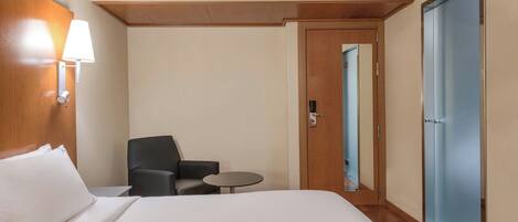 A hotel room with a bed, a chair, a small table, a lamp, and a door.