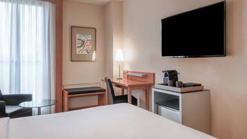 Habitación de hotel con cama, escritorio, silla, televisor, lámpara y cafetera.
