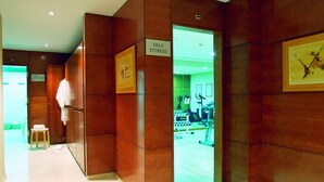 Un couloir aux murs en bois, une enseigne indiquant « Sala Fitness », et un aperçu d’une salle de sport équipée de matériel d’exercice.