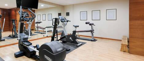 A gym with various exercise machines, a mirror, and a wooden floor.