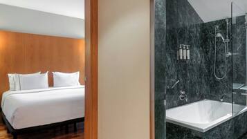 A modern hotel room with a large bed, a wooden headboard, and a marble bathroom with a bathtub and shower.