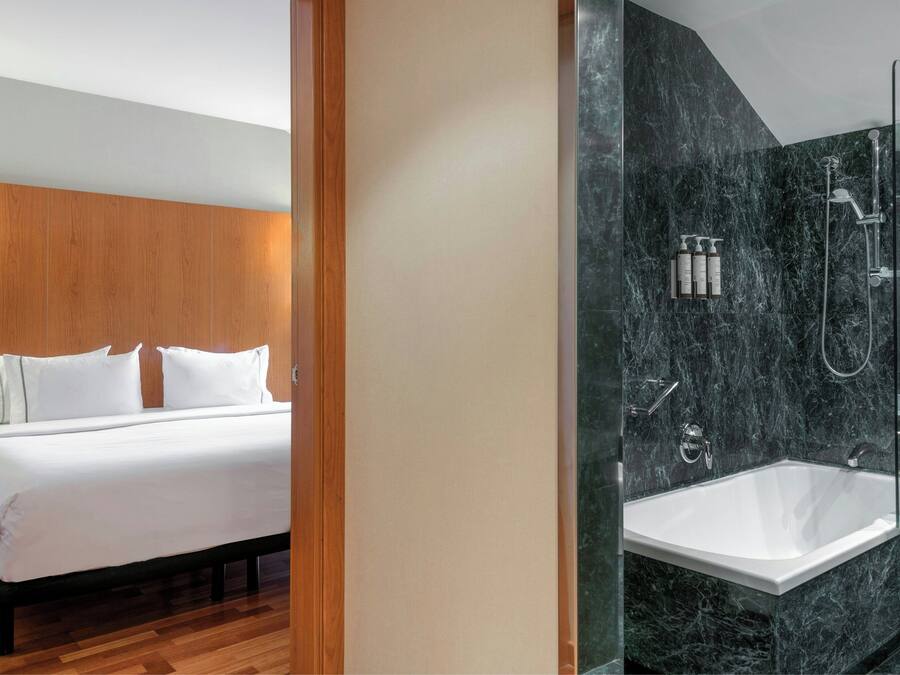 A modern hotel room with a large bed, a wooden headboard, and a marble bathroom with a bathtub and shower.