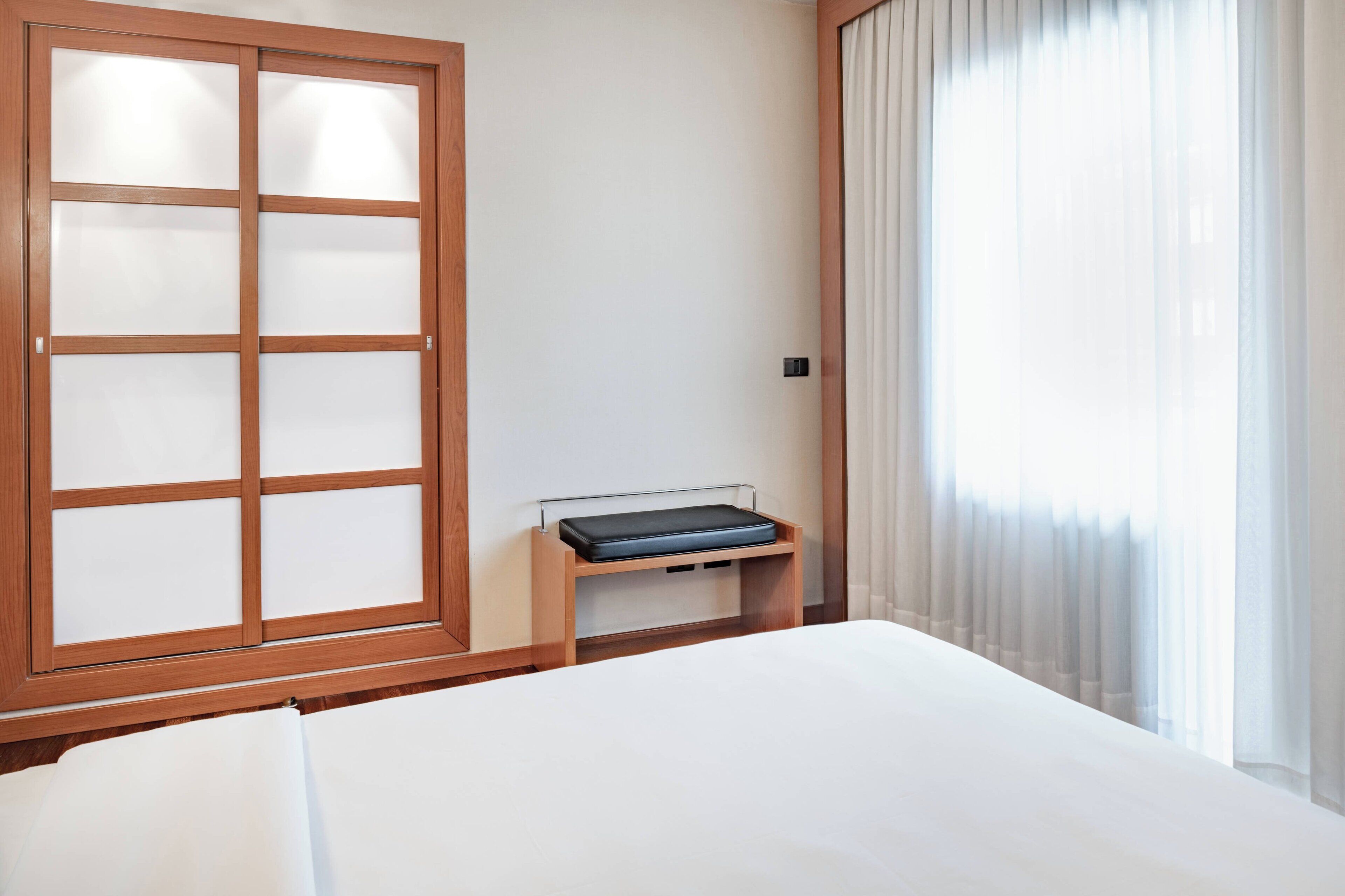 A hotel room with a large bed, a wooden cabinet with frosted glass doors, and a window with sheer curtains.