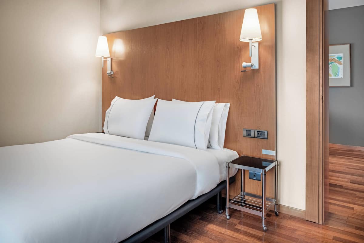 A hotel room with a large bed, two wall-mounted lamps, a small bedside table, and a wooden headboard.
