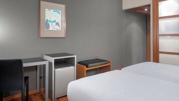 A hotel room with a bed, a desk, a chair, a small refrigerator, and a framed picture on the wall.