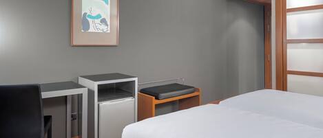 Una habitación de hotel con una cama, un escritorio, una silla, un refrigerador pequeño y un cuadro en la pared.