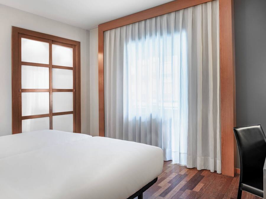 Una habitación de hotel con una cama, una ventana con cortinas, una puerta de madera y una silla.
