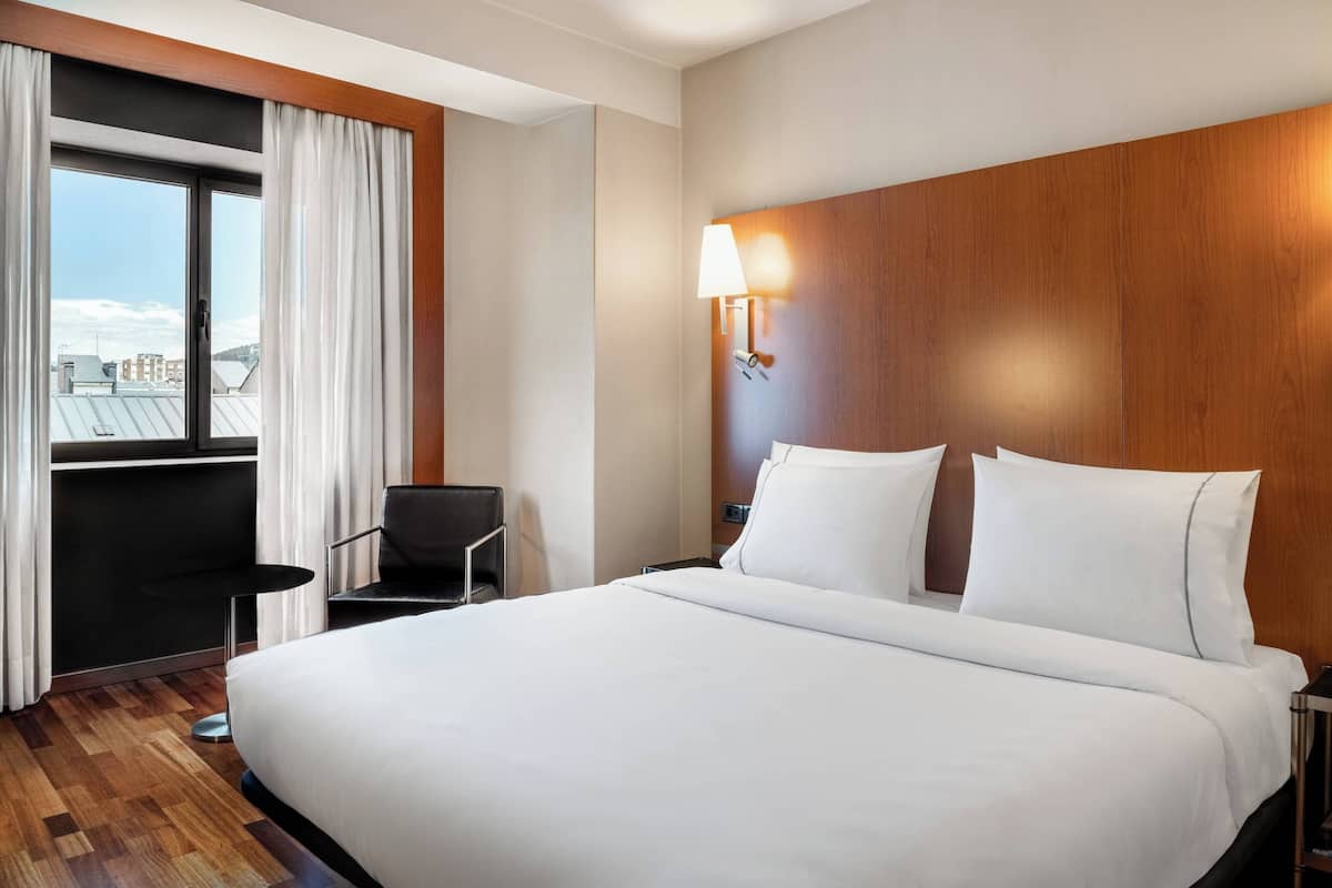 A hotel room with a large bed, a desk, a chair, a window with curtains, and a wall-mounted lamp.