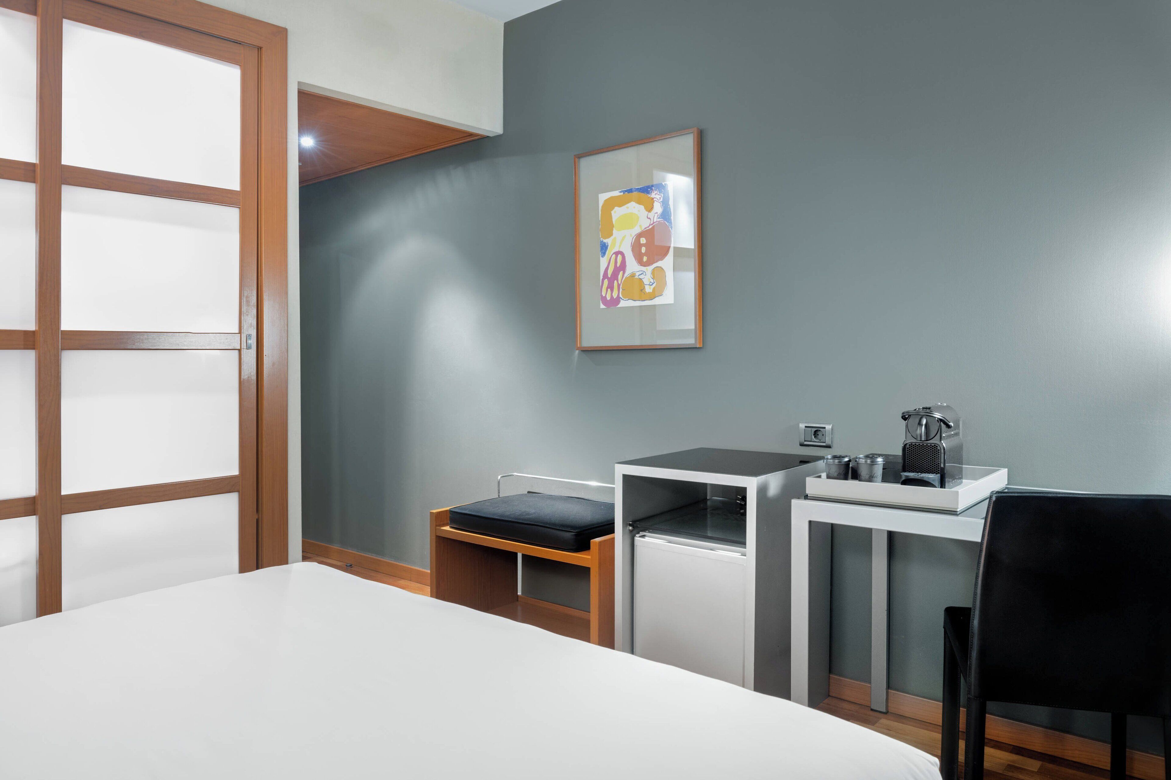 A hotel room with a bed, a desk with a chair, a small table with a kettle, and a framed picture on the wall.