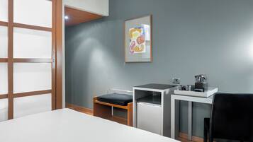 A hotel room with a bed, a desk with a chair, a small table with a kettle, and a framed picture on the wall.