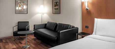 Habitación de hotel con una cama, un sofá de cuero negro, una mesita redonda pequeña, dos sillas, un cabecero de madera y dos lámparas de pared.