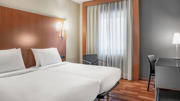 Habitación de hotel con dos camas, un escritorio, una silla, una ventana con cortinas y una lámpara fijada a la pared.
