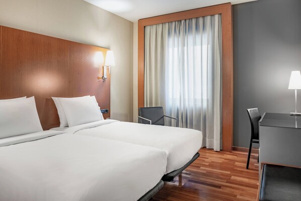 A hotel room with two beds, a desk, a chair, a window with curtains, and a wall-mounted lamp.