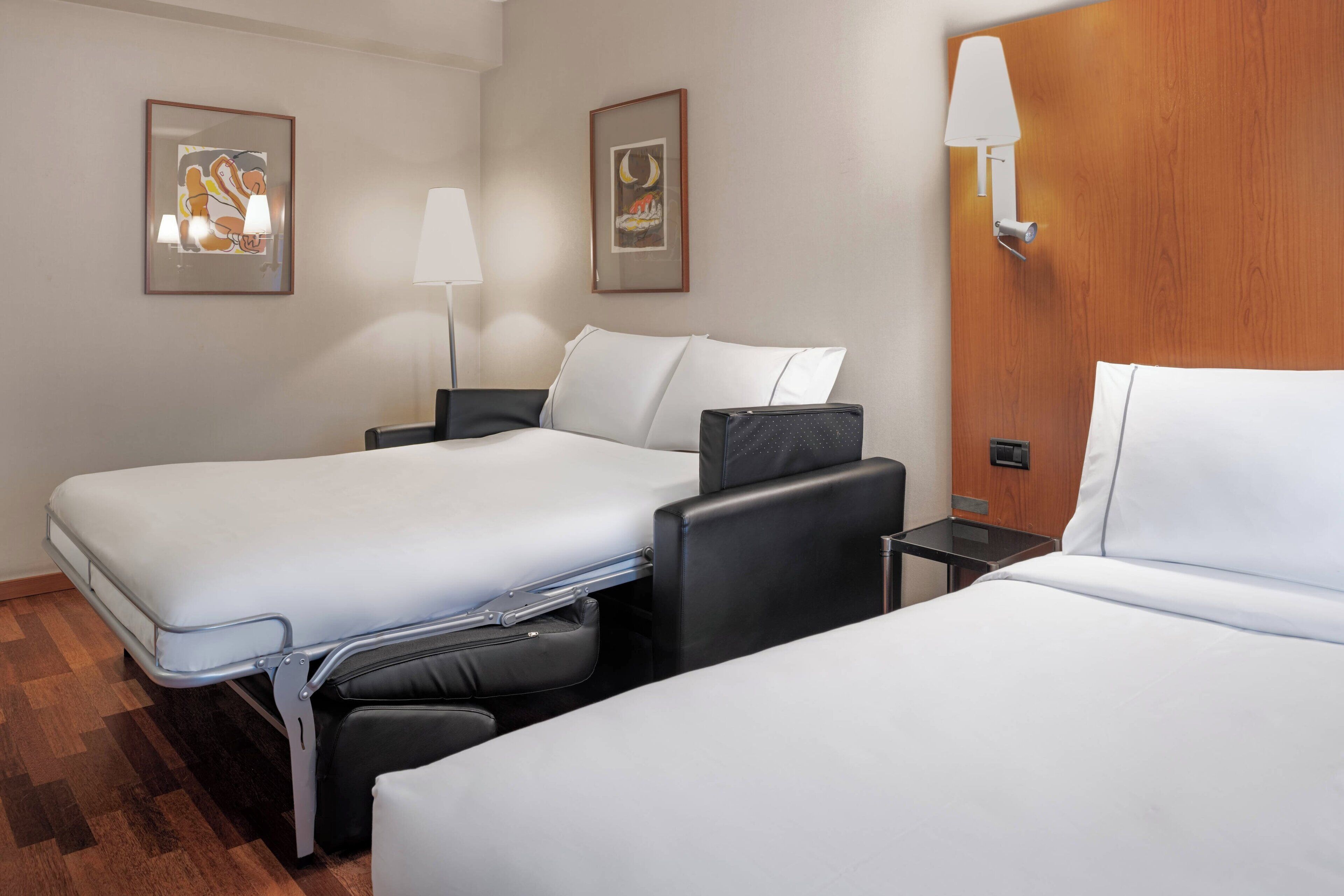 Habitació d'hotel amb dos llits, una taula de treball, una cadira, una làmpada i dos quadres penjats a la paret.