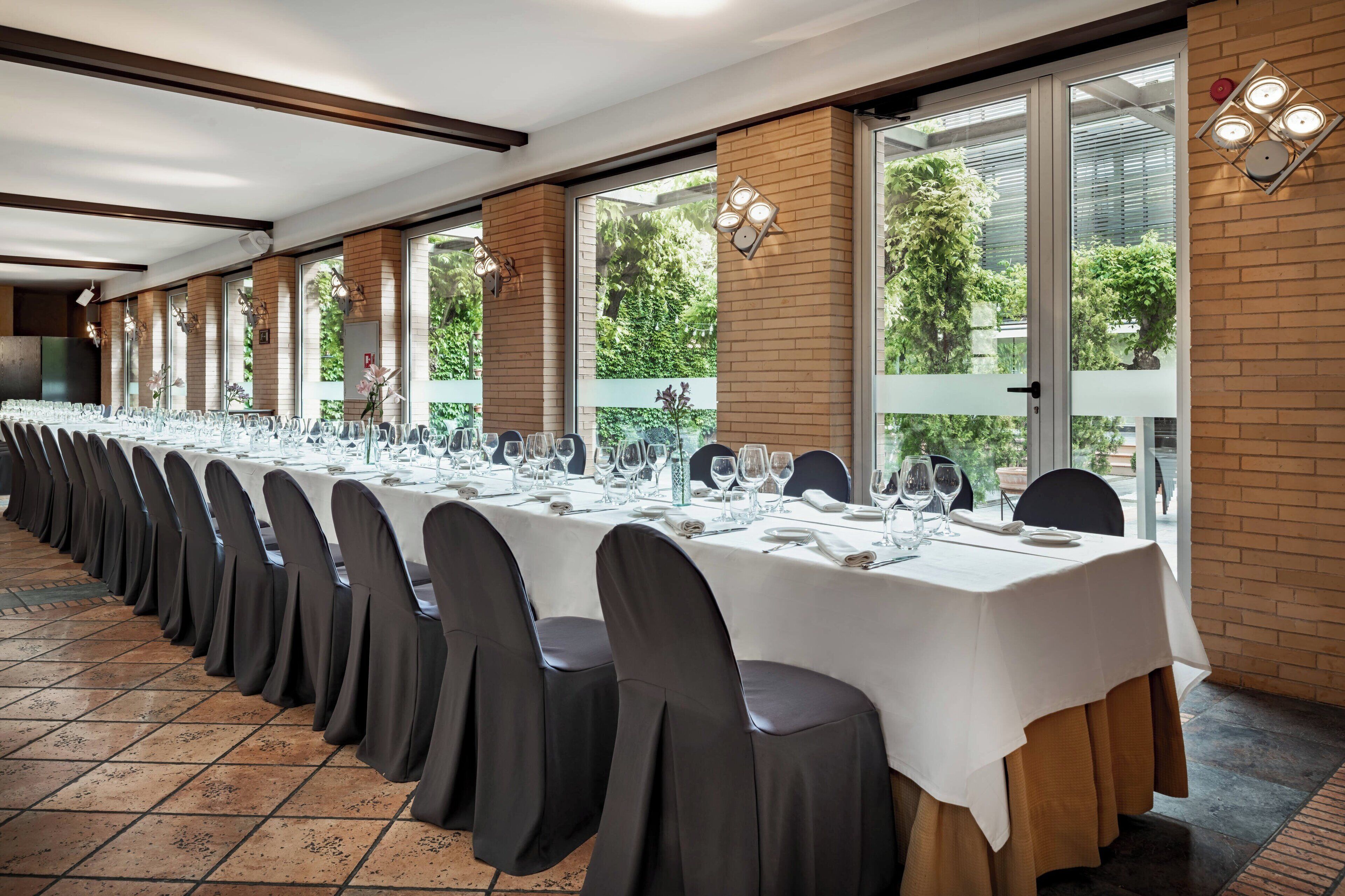 A long banquet table set with chairs, white tablecloths, and glassware, with large windows and brick walls.