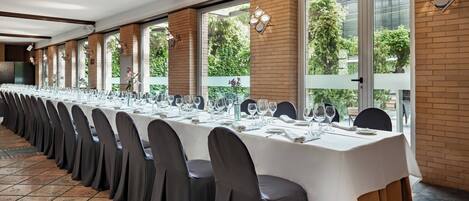 A long banquet table set with chairs, white tablecloths, and glassware, with large windows and brick walls.