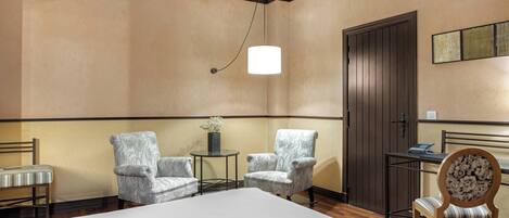 A hotel room with a large bed, two armchairs, a small table with a vase, a wooden chair, and a door.