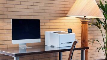 A desktop computer, a printer, a lamp, and a chair against a brick wall.