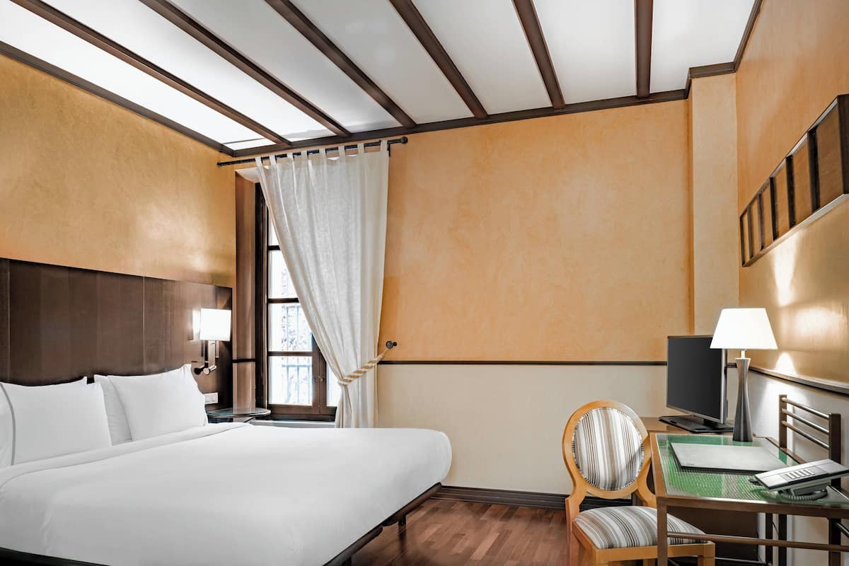 Quarto de hotel com uma cama grande, uma escrivaninha com cadeira, televisão e uma janela com cortinas.