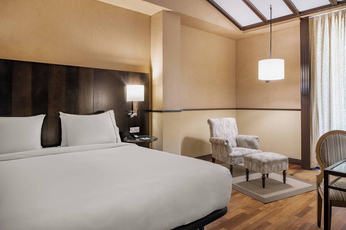 Quarto de hotel com uma cama grande, mesa de cabeceira com abajur, uma cadeira e uma mesa.