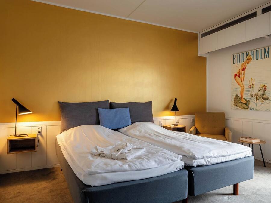 Una cama doble con sábanas blancas, dos mesitas de noche con lámparas, una silla y una pared con un cartel.