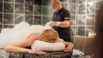 Un entorno de spa con una persona acostada en una camilla de tratamiento, otra persona atendiendo y una pared de azulejos al fondo.
