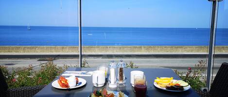 A table with breakfast items and a glass of juice, overlooking a coastal road with a view of the sea.
