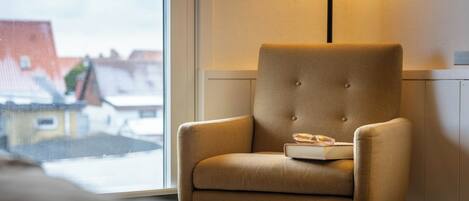 A chair with a book and glasses, a lamp, and a view of buildings through a window.