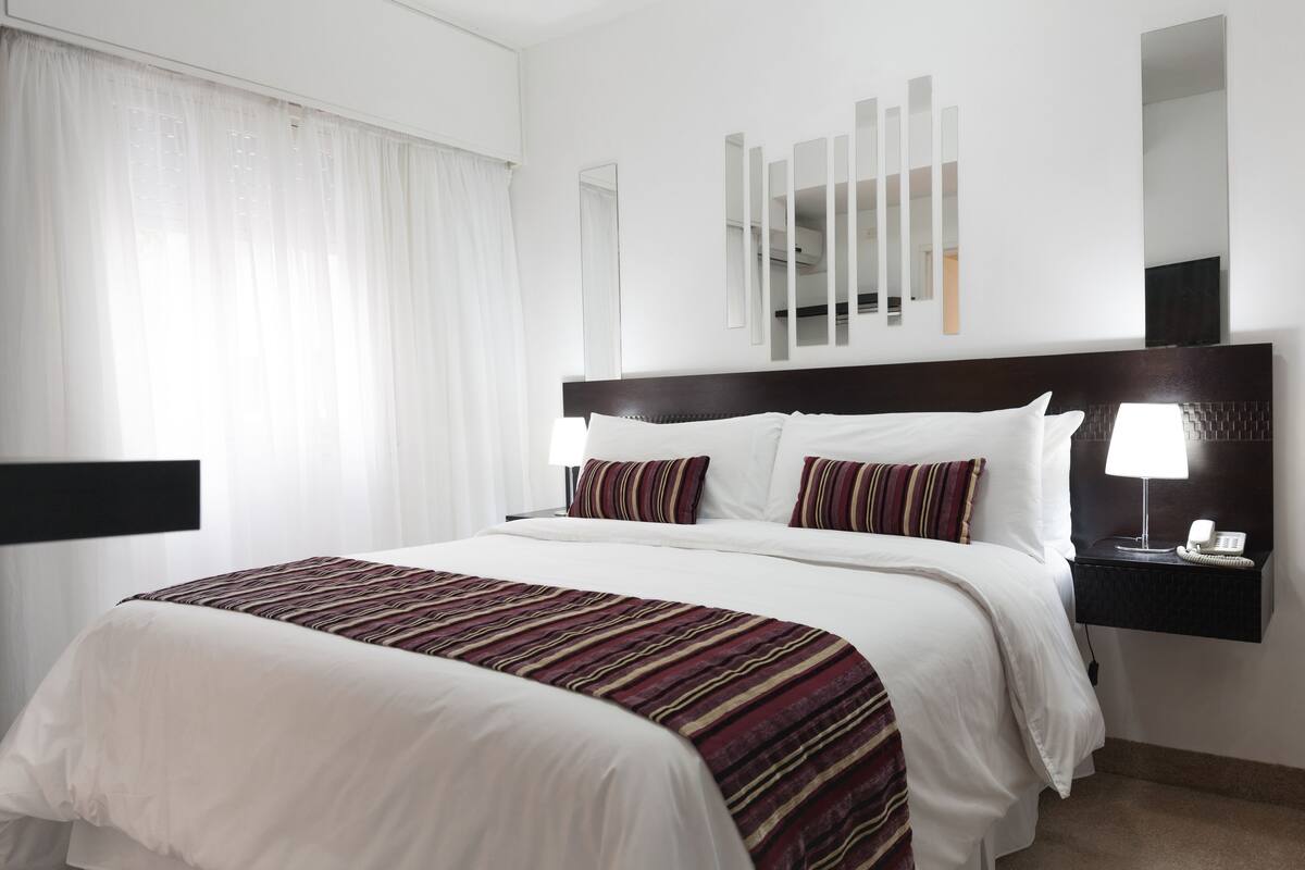 A hotel room with a large bed, white bedding, a striped throw blanket, a bedside table with a lamp, and a mirror on the wall.