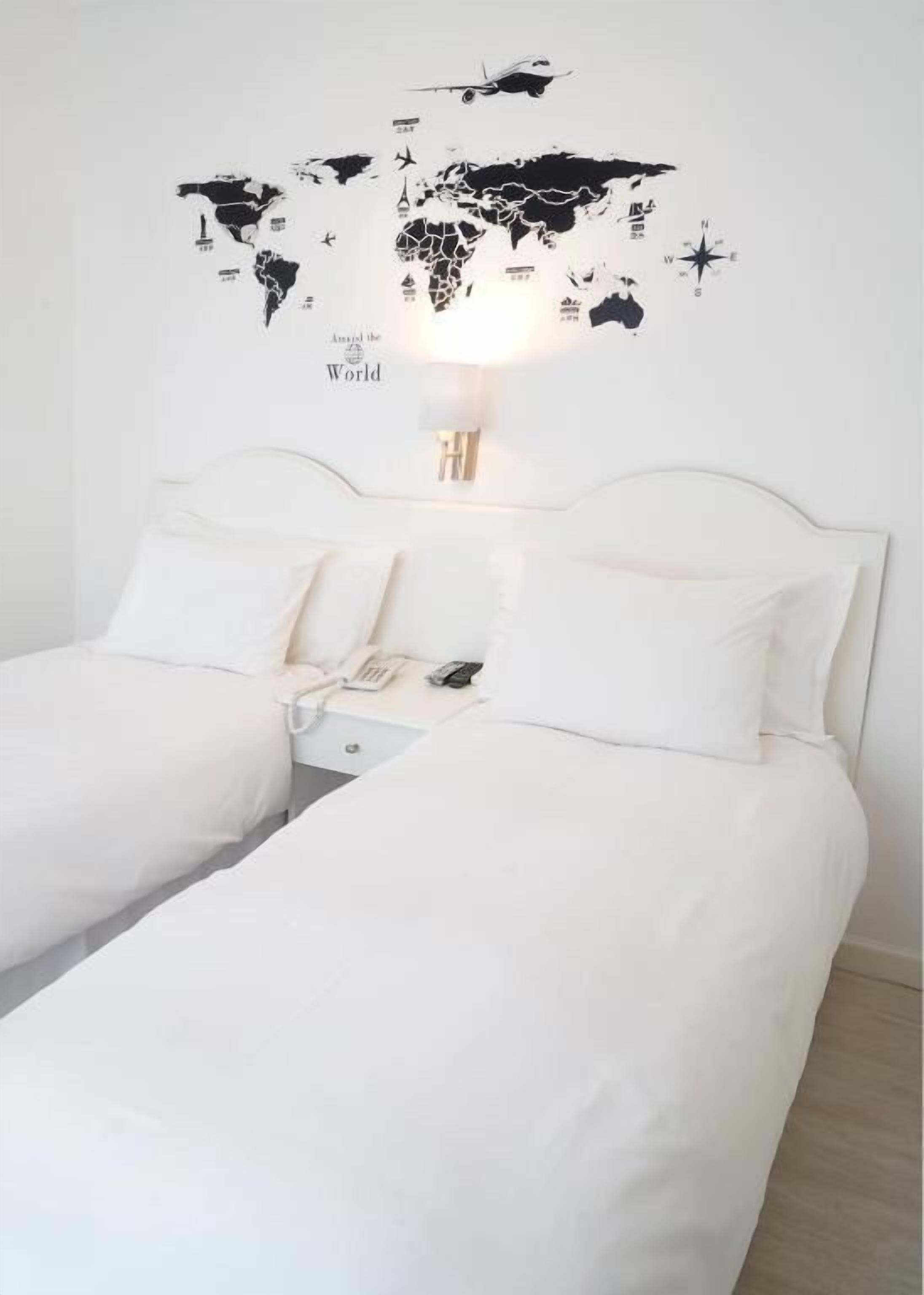 A hotel room with two beds, a world map wall art, a lamp, and a nightstand with a phone and remote.