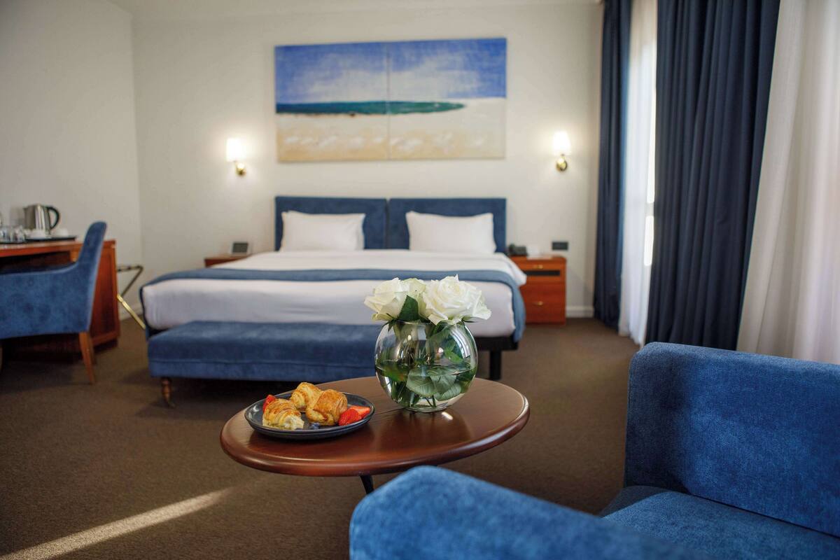 A hotel room with a bed, a desk, a chair, a table with food, and a vase with flowers.