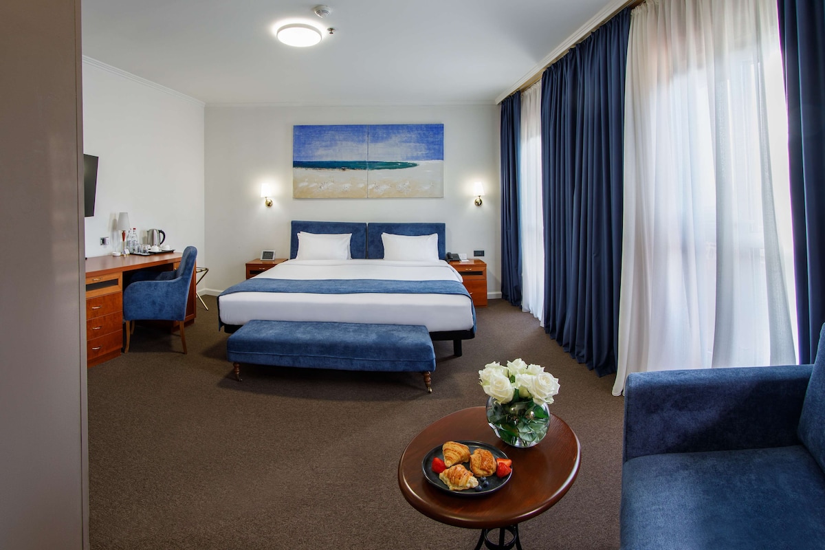 A hotel room with a bed, a desk, a chair, a coffee table with flowers, and a view of the ocean through the window.