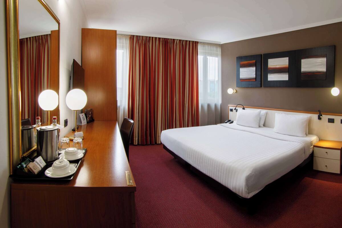 A hotel room with a bed, bedside table, lamp, mirror, and a wall with three framed pictures.