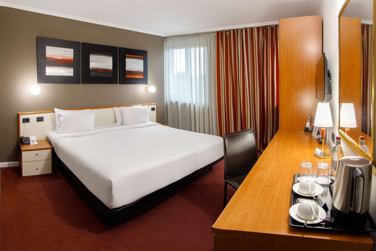 A hotel room with a large bed, a desk with a lamp, a chair, and a mirror. There are three framed pictures on the wall and a window with curtains.