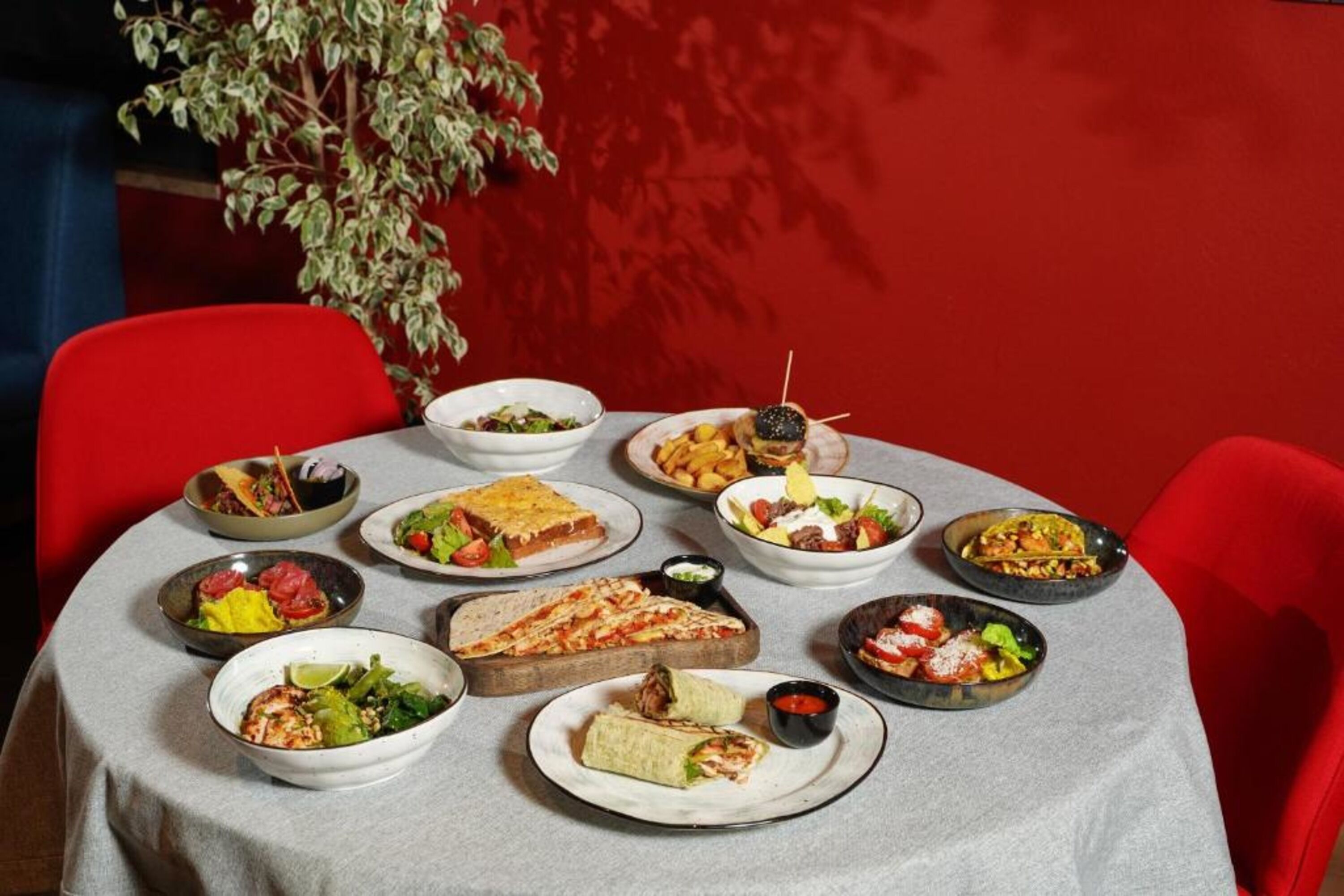 A table set with various dishes, including pizza, salads, and a burrito, in a room with red chairs and a plant.