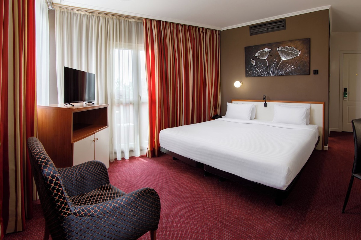 A hotel room with a bed, a desk, a chair, a television, and a painting on the wall.